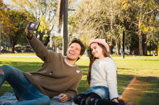 Bemannen Sie das Sitzen im Park mit seiner Tochter, die selfie mit intelligentem Telefon nimmt