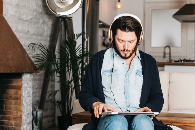 Bemannen Sie das Sitzen in der Küche unter Verwendung des Laptops, um Musik auf Kopfhörer zu hören