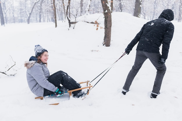 Bemannen Sie das Ziehen des lächelnden Mädchens auf Schlitten durch schneebedeckte Landschaft