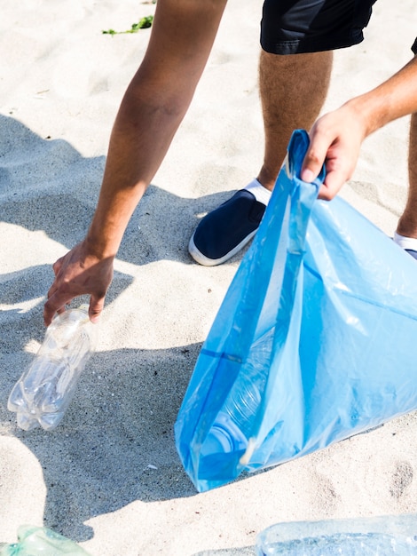 Bemannen Sie die Hand, die Plastikflasche beim Halten der blauen Abfalltasche auf Strand auswählt