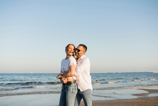 Bemannen Sie die Umfassung seiner Freundin von hinten, die nahe dem Meer gegen blauen Himmel am Strand steht