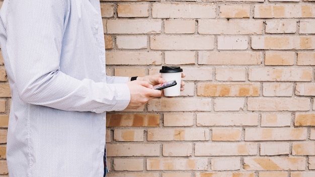 Bemannen Sie Stellung vor Backsteinmauer mit der Kaffeetasse und Handy, die in der Hand halten