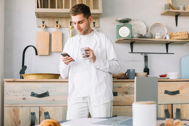 Bemannen Sie Stellung vor Küchenarbeitsplatte unter Verwendung des Handys, der Kaffeetasse hält