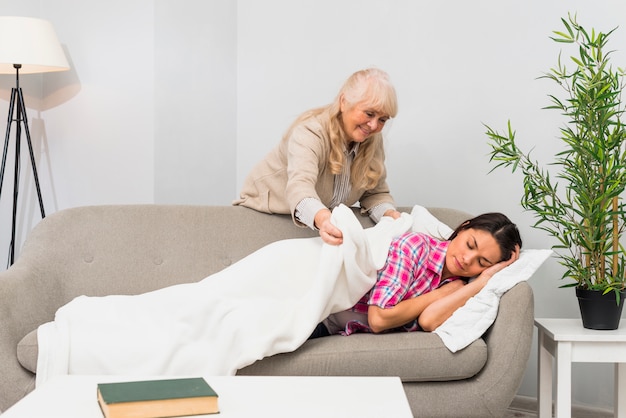 Bemuttern Sie das Setzen der Decke über ihrer jungen erwachsenen Tochter, die auf Sofa schläft