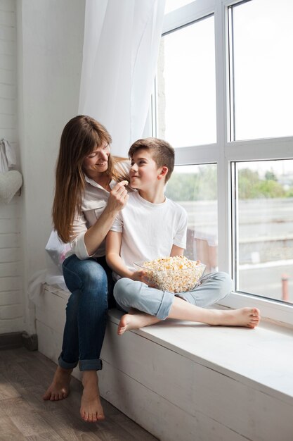 Bemuttern Sie Fütterungspopcorn zu ihrem Sohn, der zu Hause nahe Fensterbrett sitzt