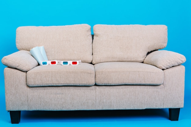 Bequeme Couch mit 3D-Brille, Popcorn lokalisiert auf blauem Hintergrund. Vorbereitung zum Ansehen von Filmen, Entspannung, Genießen des Kinos zu Hause