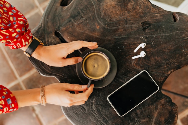 Über dem Rahmen der Frauenhand, die eine Tasse Kaffee mit Kopfhörern und Smartphone hält