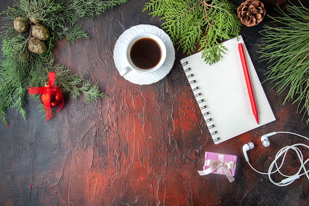 Über der Ansicht von Tannenzweigen eine Tasse schwarzen Tee Dekorationszubehör weißer Kopfhörer und Geschenk neben Notizbuch mit Stift auf dunklem Hintergrund