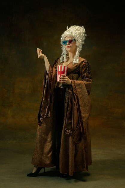 Bereit für das Kino. Porträt der mittelalterlichen Frau in der Weinlesekleidung mit 3D-Brille, Popcorn auf dunklem Hintergrund. Weibliches Modell als Herzogin, königliche Person. Konzept des Vergleichs von Epochen, Mode, Schönheit.
