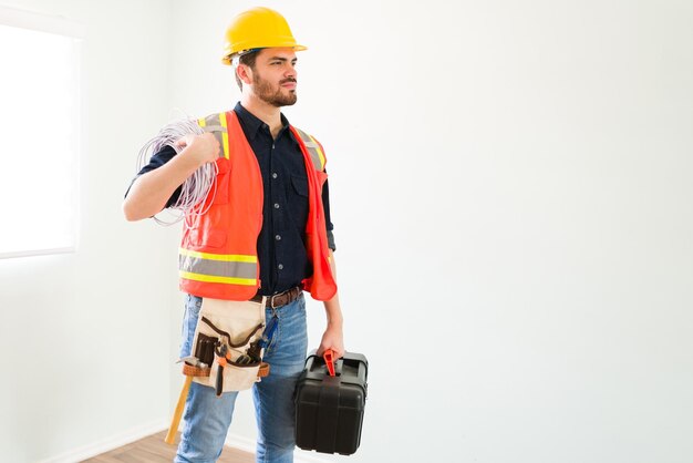 Bereit, meine Arbeit zu beginnen. Stolzer attraktiver Elektriker mit Schutzausrüstung, der einen Werkzeugkasten und Stromkabel hält