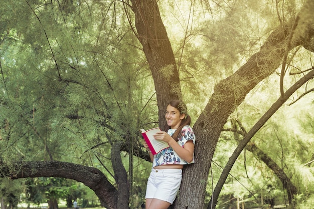 Kostenloses Foto bereitstehender baum des mädchens, der das buchlächeln hält