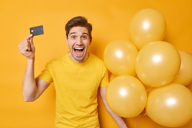 Überemotionaler fröhlicher junger Mann im lässigen T-Shirt hält Kreditkarte und einen Haufen Heliumballons bereit zum Feiern, um alles Geld isoliert auf gelbem Hintergrund auszugeben, feiert Geburtstag