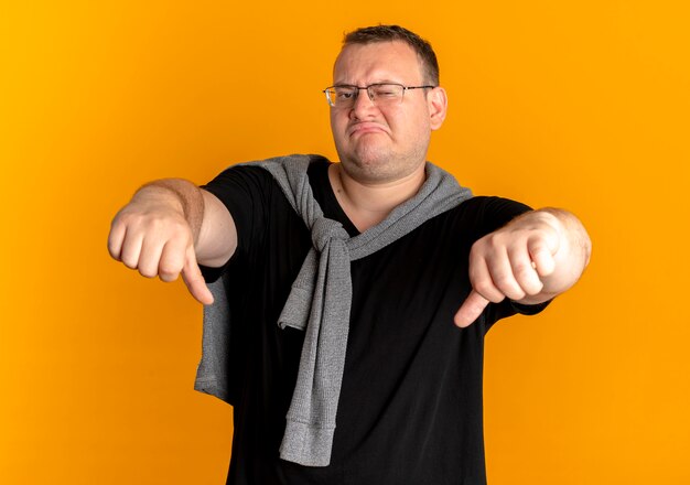 Übergewichtiger Mann in Brille, der schwarzes T-Shirt trägt, missfiel, Daumen über Orange zeigend