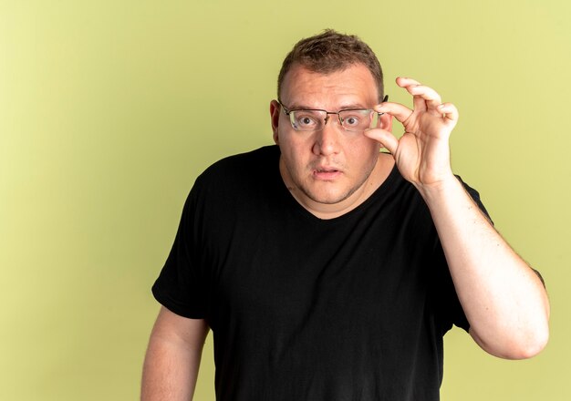 Übergewichtiger Mann in Brille mit schwarzem T-Shirt überraschte, als er seine Brille über einer hellen Wand befestigte
