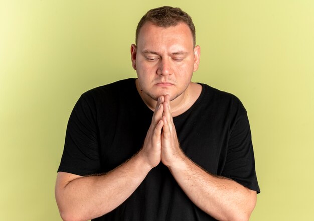 Übergewichtiger Mann in der Brille, der schwarzes T-Shirt trägt, das Hände zusammenhält, wie mit Hoffnungsausdruck über Licht zu beten