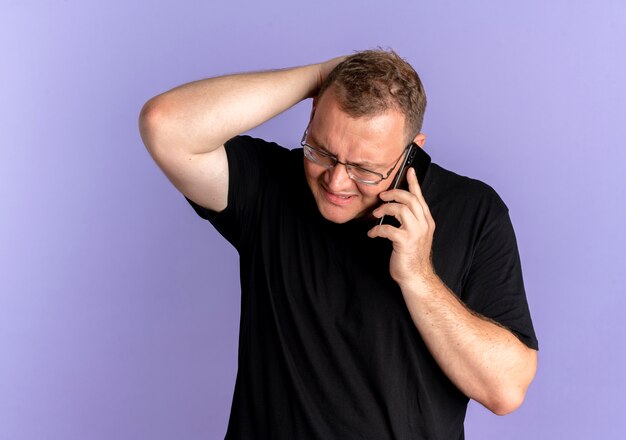 Übergewichtiger Mann in der Brille, der schwarzes T-Shirt trägt, das verwirrt und unzufrieden aussieht, während auf Handy über Blau spricht