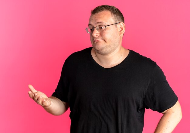 Übergewichtiger Mann in der Brille, die das schwarze T-Shirt trägt, das beiseite schaut, mit dem Arm heraus denkend oder fragend steht über rosa Wand