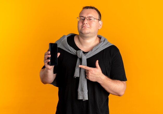 Übergewichtiger Mann in der Brille, die das schwarze T-Shirt trägt, das Smartphone zeigt, das mit dem Finger darauf zeigt, der über Orange lächelt