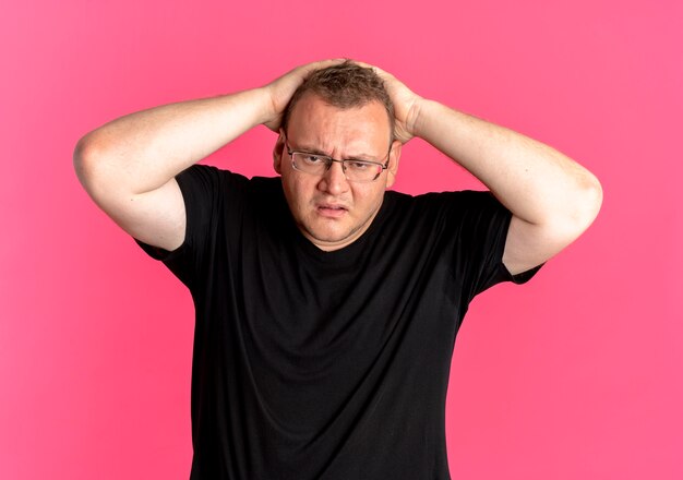 Übergewichtiger Mann in der Brille, die das schwarze T-Shirt trägt, das verwirrt schaut und seinen Kopf mit den Händen über rosa hält