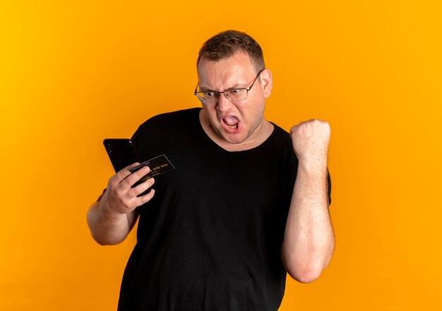 Übergewichtiger Mann in der Brille, die schwarzes T-Shirt hält, das die geballte Faust des Smartphones hält, die über Orange schreit