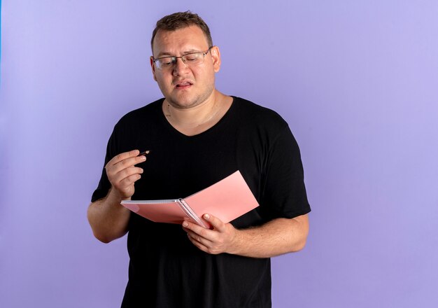 Übergewichtiger Mann in der Brille, die schwarzes T-Shirt hält, das Notizbuch und Stift hält, die verwirrt stehen über blauer Wand stehen