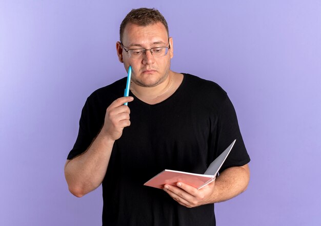 Übergewichtiger Mann in der Brille, die schwarzes T-Shirt hält, das Notizbuch und Stift hält und mit nachdenklichem Ausdruck über blauer Wand steht