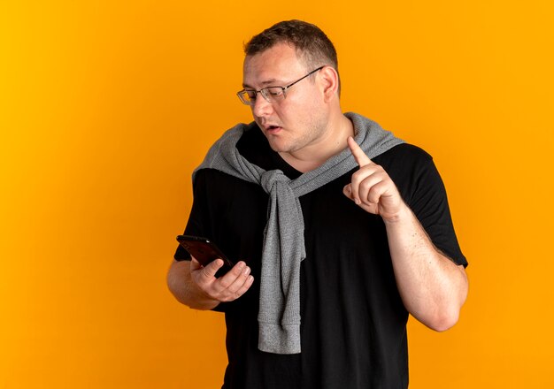 Übergewichtiger Mann in der Brille, die schwarzes T-Shirt hält, das Smartphone hält, das verwirrt zeigt Zeigefinger über Orange zeigt