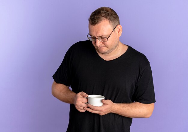 Übergewichtiger Mann in der Brille, die schwarzes T-Shirt hält Kaffeetasse hält, die es mit nachdenklichem Ausdruck über Blau betrachtet