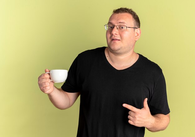Übergewichtiger Mann in der Brille, die schwarzes T-Shirt hält Kaffeetasse hält, die mit dem Finger darauf zeigt, mit glücklichem Gesicht, das über Lichtwand steht