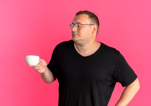 Übergewichtiger Mann in der Brille, die schwarzes T-Shirt hält Kaffeetasse hält, die mit ernstem Gesicht über rosa beiseite schaut