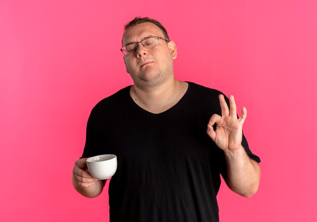 Übergewichtiger Mann in der Brille, die schwarzes T-Shirt hält Kaffeetasse hält ok Zeichen erfreut über rosa Wand stehen