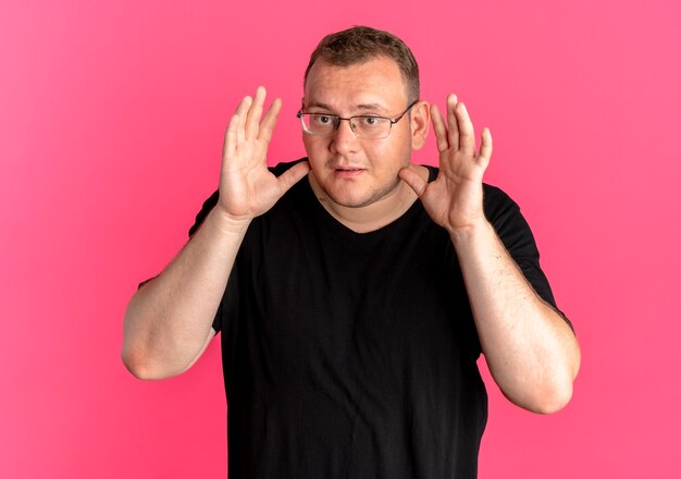 Übergewichtiger Mann in der Brille, die schwarzes T-Shirt mit Armen nahe Gesicht über rosa trägt