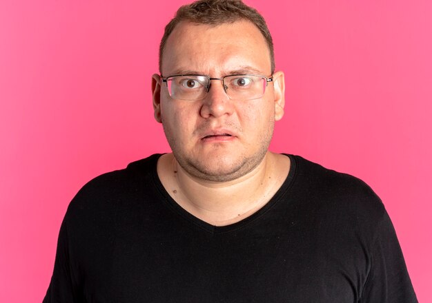 Übergewichtiger Mann in der Brille, die schwarzes T-Shirt mit Blick auf die Kamera trägt, überrascht und unzufrieden, über rosa Wand stehend