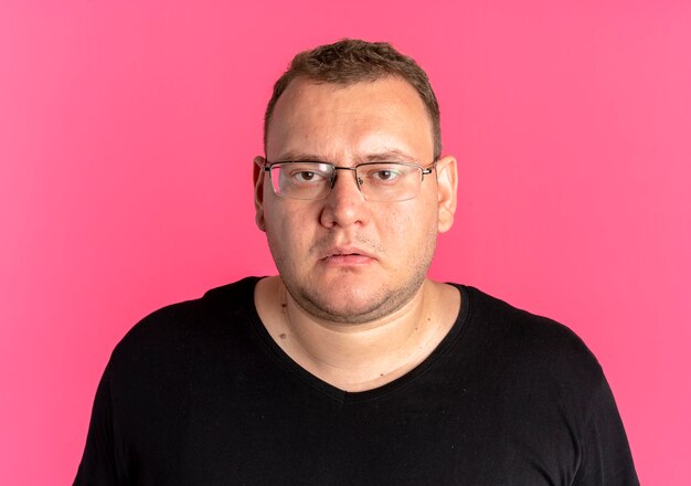 Übergewichtiger Mann in der Brille, die schwarzes T-Shirt mit ernstem Gesicht über rosa trägt