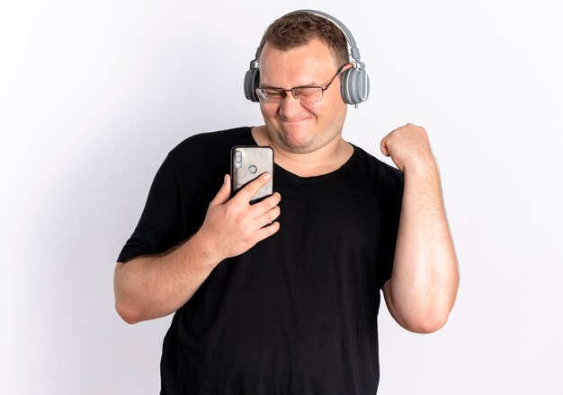 Übergewichtiger Mann in der Brille, die schwarzes T-Shirt mit Kopfhörern hält, die Smartphone halten, das seine Lieblingsmusik genießt, die über weißer Wand steht