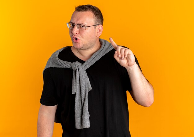 Übergewichtiger Mann in der Brille, die schwarzes T-Shirt mit stirnrunzelndem Gesicht trägt, das Zeigefinger über Orange zeigt