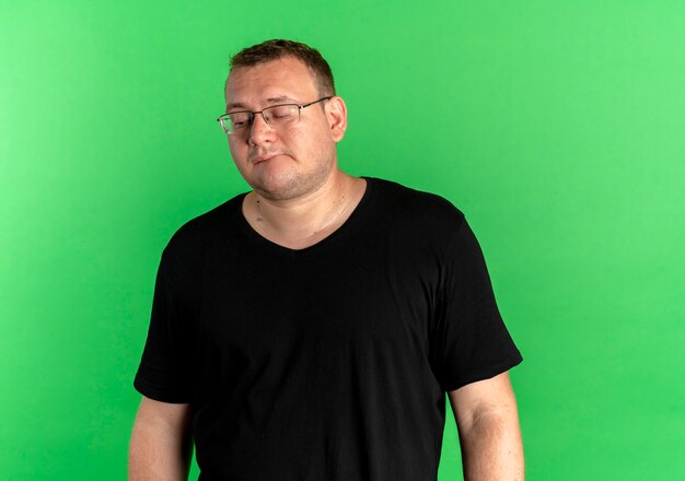 Übergewichtiger Mann in der Brille, die schwarzes T-Shirt mit traurigem Ausdruck auf Gesicht steht, das über grüner Wand steht