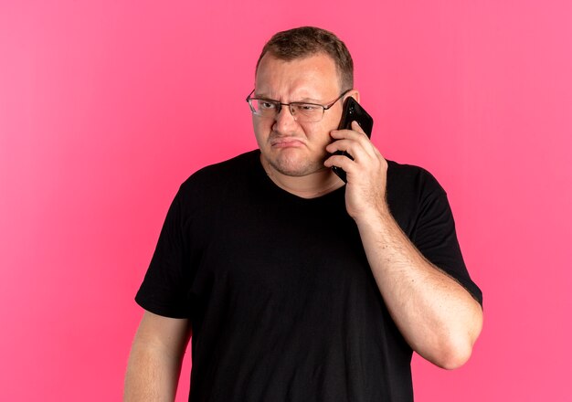 Übergewichtiger Mann in der Brille, die schwarzes T-Shirt trägt, das beim Sprechen über Handy über Rosa unzufrieden aussieht