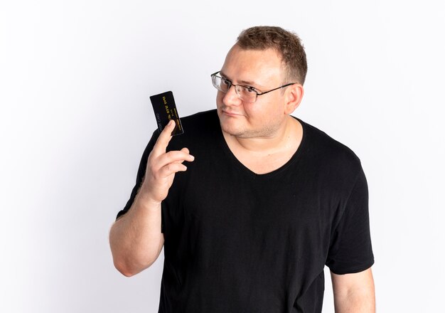 Übergewichtiger Mann in der Brille, die schwarzes T-Shirt trägt, das Kreditkarte betrachtet, die Kamera verwirrt über weißer Wand stehend betrachtet
