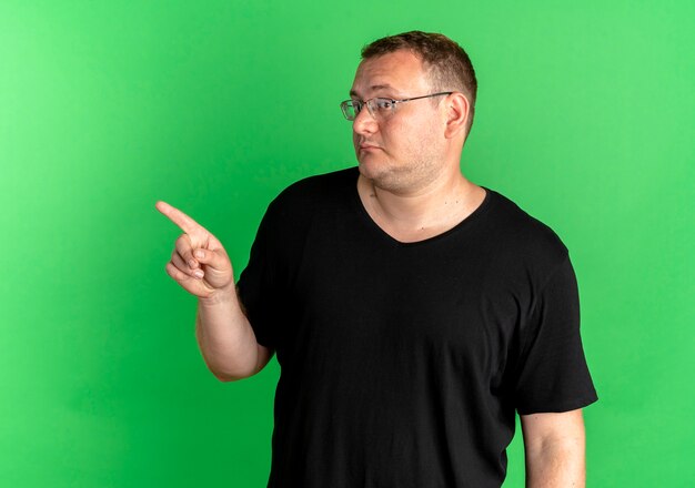 Übergewichtiger Mann in der Brille, die schwarzes T-Shirt trägt, das mit dem Finger zur Seite zeigt, verwirrt über Grün schaut