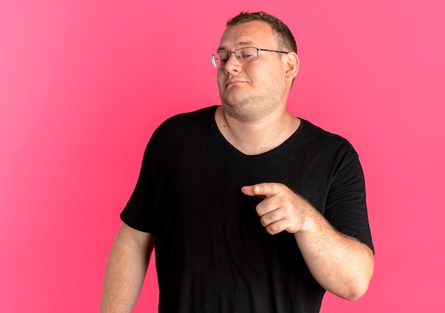 Übergewichtiger Mann in der Brille, die schwarzes T-Shirt trägt, das mit Zeigefinger zur Seite über Rosa zuversichtlich zeigt