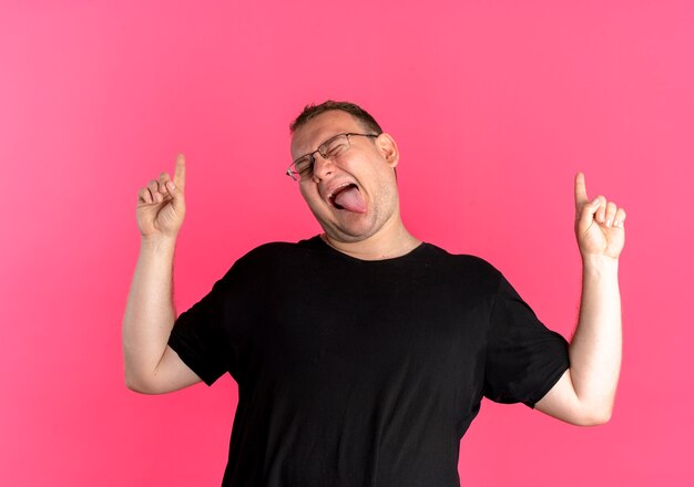 Übergewichtiger Mann in der Brille, die schwarzes T-Shirt trägt, das Spaß hat, herauszungende Zunge heraus zu zeigen, die mit Zeigefingern zeigt, die über rosa Wand stehen
