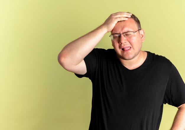 Übergewichtiger Mann in der Brille, die schwarzes T-Shirt trägt, das verwirrt mit der Hand auf seinem Kopf für Fehler steht, der über leichter Wand steht