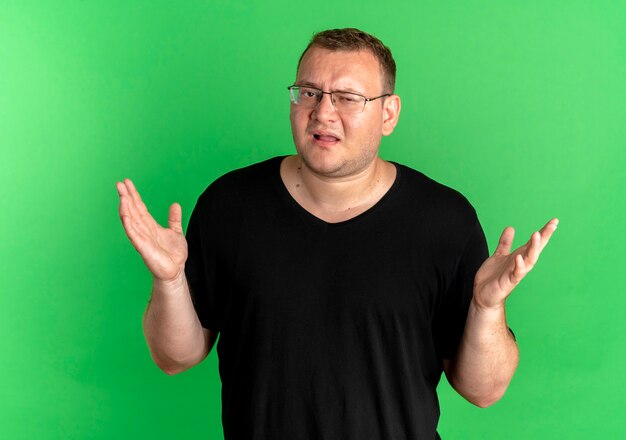 Übergewichtiger Mann in der Brille, die schwarzes T-Shirt trägt, das verwirrte achselzuckende Schultern über grüner Wand steht