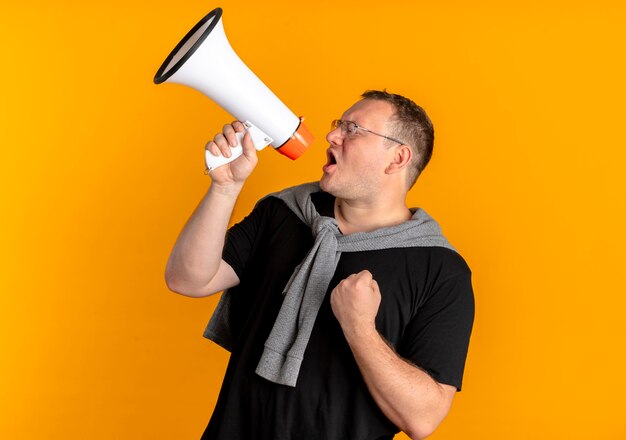 Übergewichtiger Mann in der Brille, die schwarzes T-Shirt trägt, das zu Megaphon schreit, das Faust ballt, die über orange Wand steht