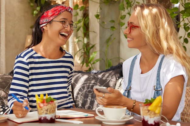 Überglücklich lachen zwei Frauen glücklich, während sie Meinungen über das Planungsprojekt austauschen, während der Kaffeepause kommunizieren, Aufzeichnungen im Veranstalter schreiben, köstliche Nachspeisen essen, Freizeitkleidung und Brillen tragen