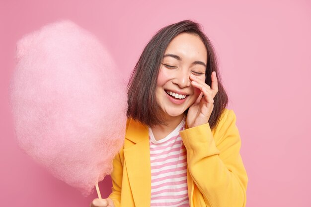 Überglückliche brünette junge asiatische Frau schließt die Augen und lächelt glücklich hat Spaß beim Gehen auf der Straße während des Sommertages hält köstliche Zuckerwatte isoliert über rosa Wand bekommt süße Leckerei