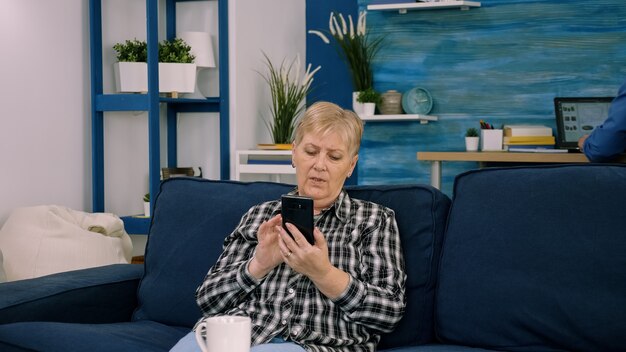 Überglückliche Frau mittleren Alters, die auf der Couch im Wohnzimmer sitzt und über einen Videoanruf auf dem Smartphone-Gadget spricht, glückliche, aufgeregte weibliche Begrüßung spricht im Web mit Smartphone, Technologiekonzept