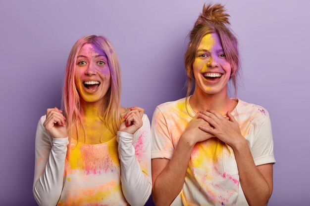 Überglückliche Frauen, die mit buntem Puder verschmiert sind, haben Spaß beim Festival of Colors in Indien, ballen die Fäuste, halten beide Handflächen auf der Brust, tragen lässige weiße Kleidung und freuen sich über den kommenden Frühling. Holi Urlaub