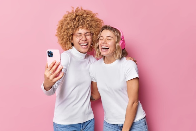 Überglückliche Frauen posieren für Selfie lachen gerne Spaß Spaß halten Smartphone vor in Freizeitkleidung gekleidet verwenden Stereo-Kopfhörer isoliert über rosa Hintergrund Zwei Freunde machen ein Foto von sich